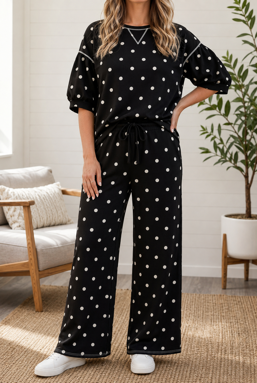 A woman modeling a black two-piece lounge set with white polka dots, featuring a short-sleeve top with puff sleeves and matching wide-leg pants, styled in a bright living room setting.