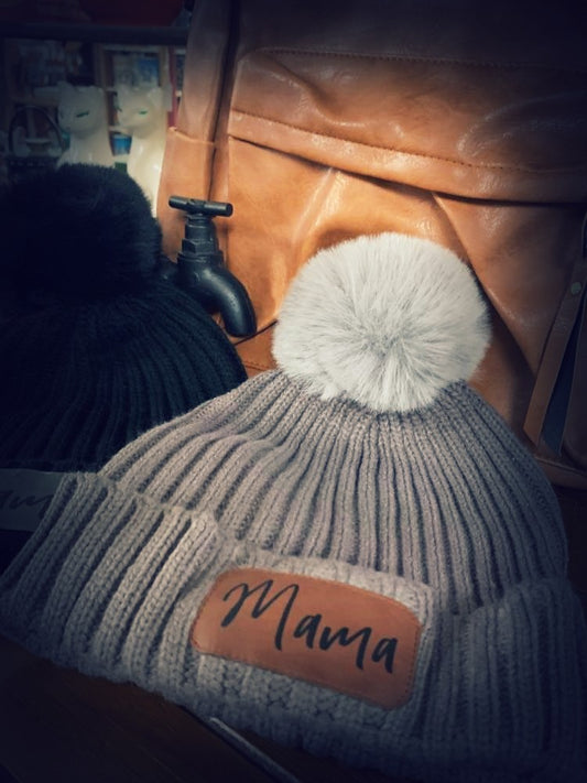Gray knit beanie with white pom-pom and 'Mama' patch on a leather surface.