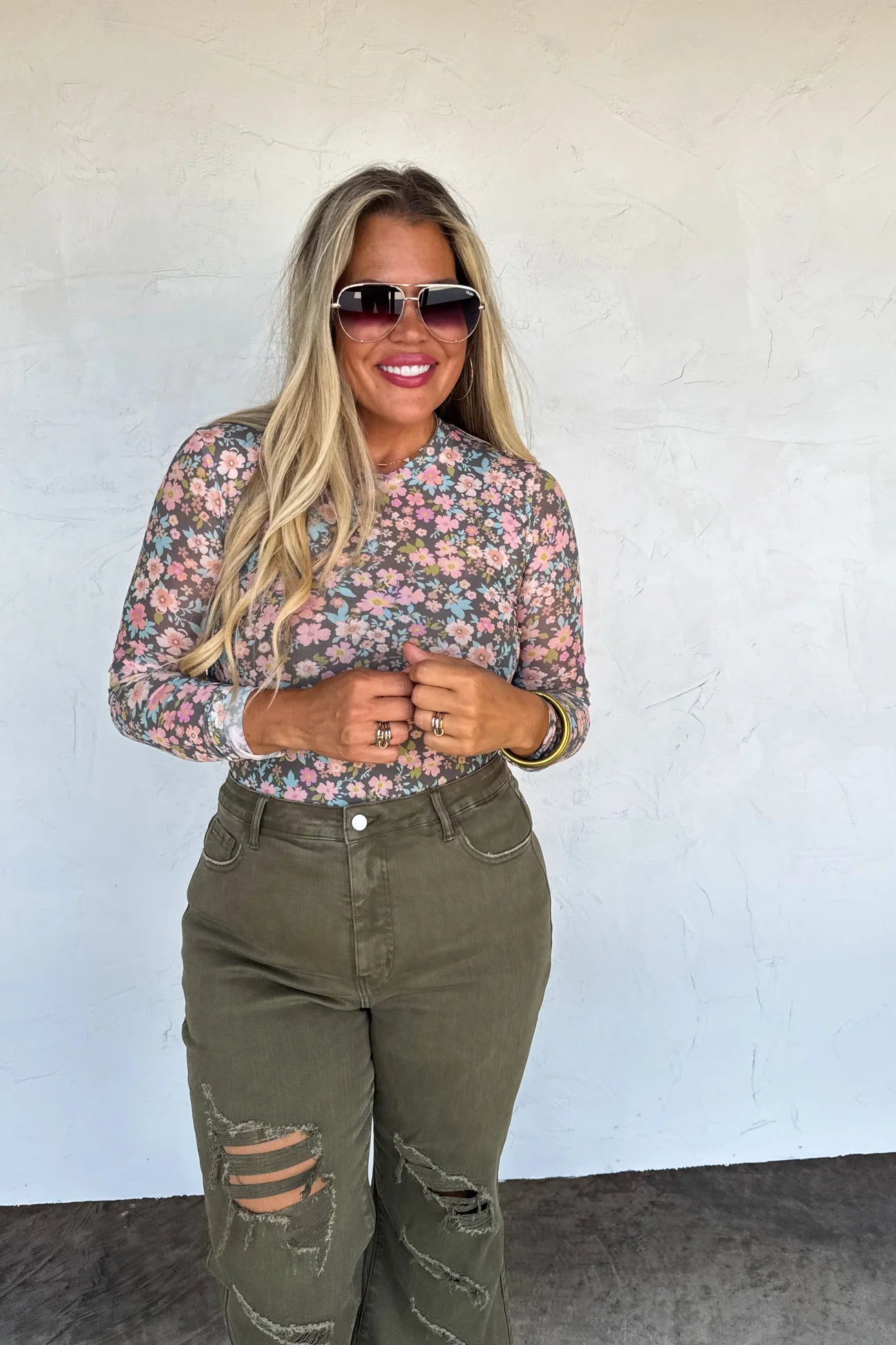 Woman wearing a floral blouse and green jeans against a white wall