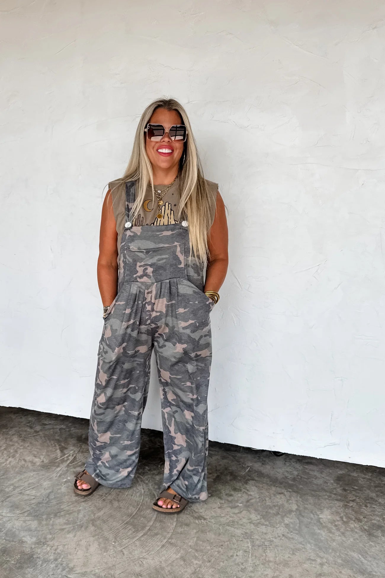 Woman wearing camouflage overalls and sunglasses against a white wall.