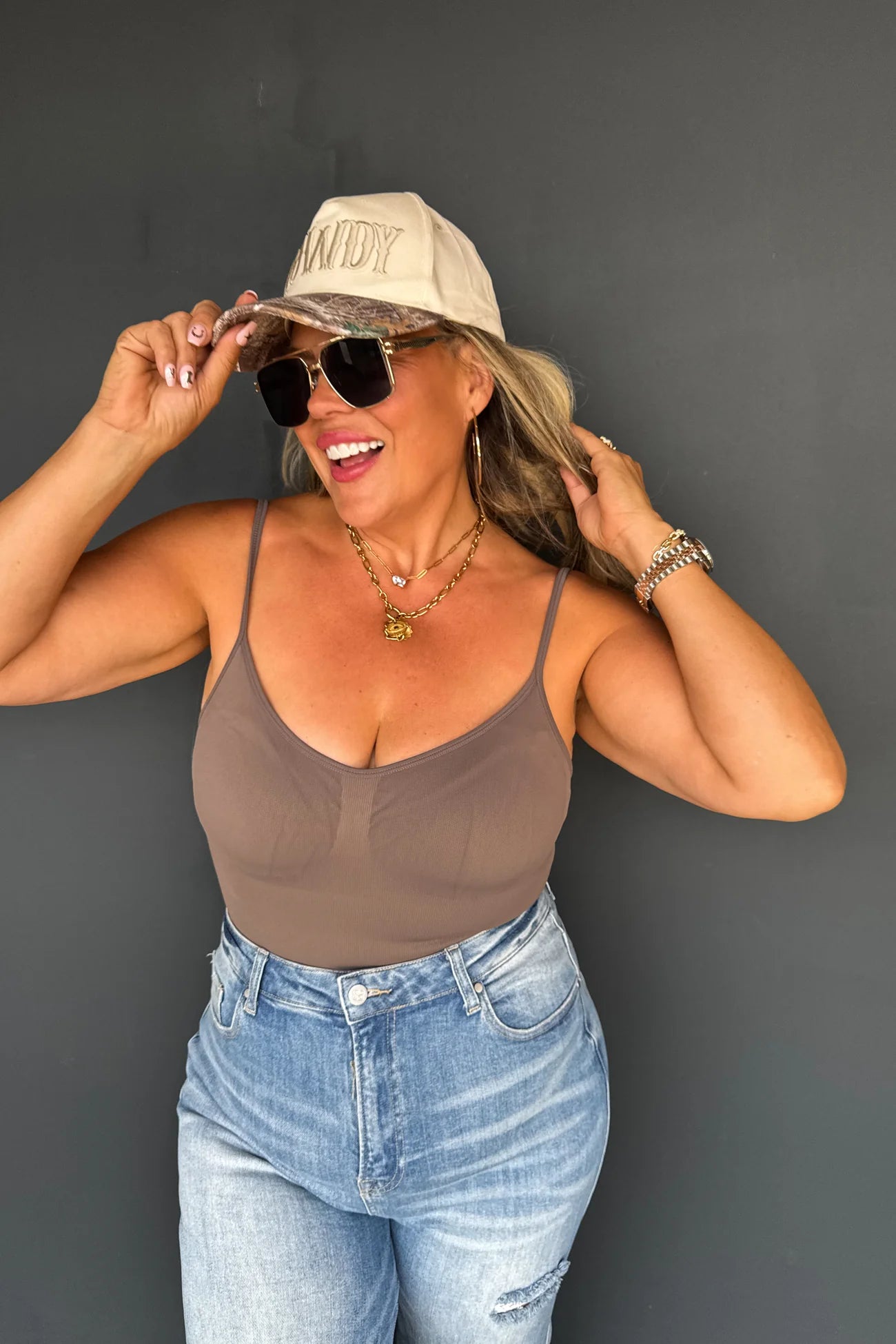 Woman wearing a beige cap, sunglasses, brown tank top, and blue jeans against a gray background