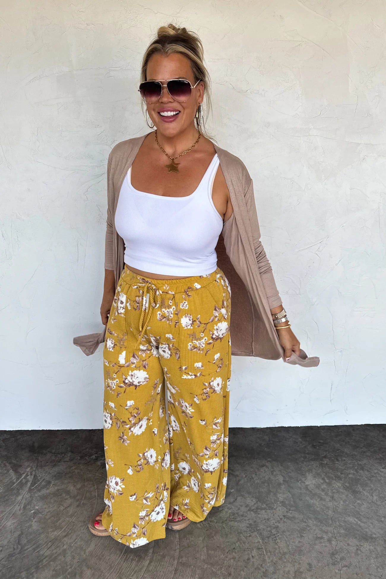 Woman wearing a white tank top, yellow floral pants, and a beige cardigan against a white wall.