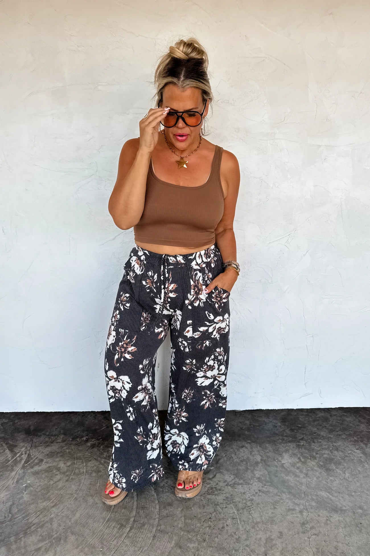 Woman wearing a brown crop top and black floral pants against a white wall.