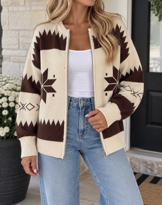 A woman wearing a cream and dark brown knit zip-up cardigan with a geometric snowflake pattern, styled over a white tank top and light blue jeans.