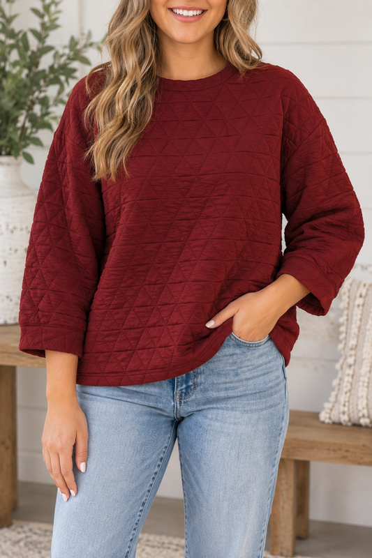 A woman wearing a textured, quilted long-sleeve sweater in deep burgundy with light wash blue jeans, standing in a brightly lit indoor setting.
