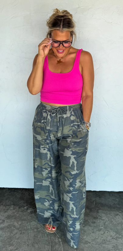 Woman wearing a pink top and camouflage pants against a white wall.